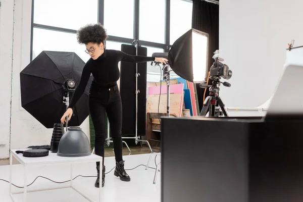 Productor de contenido afroamericano en ropa negra alcanzando lámparas cerca de reflector softbox y cámara digital en estudio fotográfico moderno - foto de stock