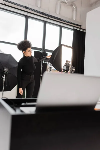 Afro-américain fabricant de contenu dans les lunettes près de réflecteur softbox dans le studio photo sur le premier plan flou — Photo de stock