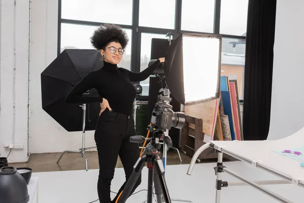 Soddisfatto produttore di contenuti africani americani in piedi con mano sul fianco vicino softbox riflettore e fotocamera digitale offuscata in studio fotografico — Foto stock