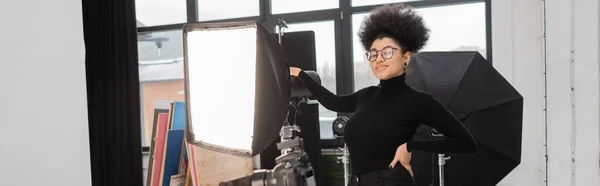 Productor de contenido afroamericano feliz posando con la mano en la cadera cerca del reflector softbox en el estudio de fotos, pancarta - foto de stock