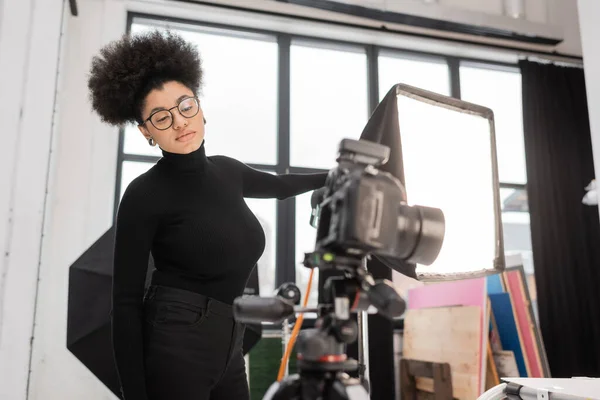 African american content maker in eyeglasses looking at blurred digital camera while standing near spotlight in photo studio — Stock Photo