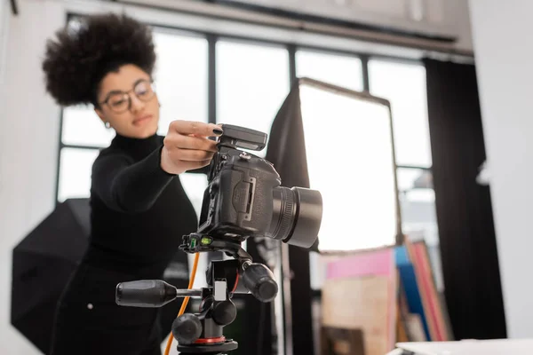 Produtor de conteúdo americano africano desfocado ajustando câmera digital perto de holofotes no estúdio de fotos — Fotografia de Stock