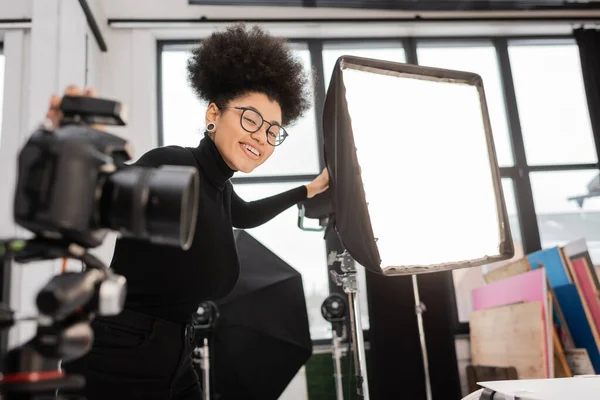 Alegre africano americano contenido fabricante sonriendo a cámara cerca reflector y borrosa cámara digital en estudio de fotos - foto de stock