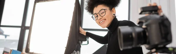 Producteur de contenu afro-américain joyeux dans des lunettes regardant la caméra près des projecteurs et appareil photo numérique flou, bannière — Photo de stock