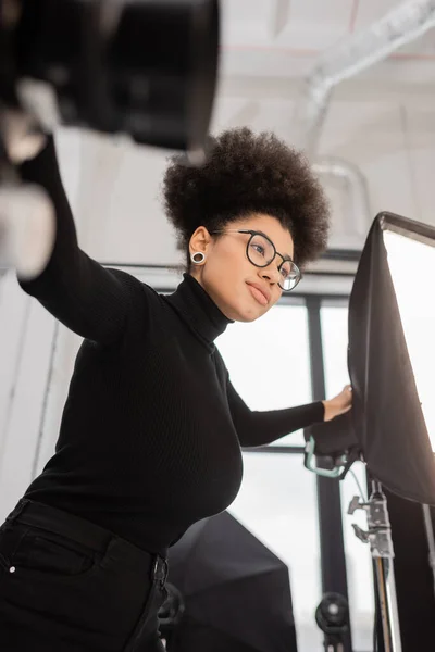 Visão de baixo ângulo de gerente de conteúdo americano africano elegante em óculos e gola alta preta perto de holofotes no estúdio de fotos — Fotografia de Stock