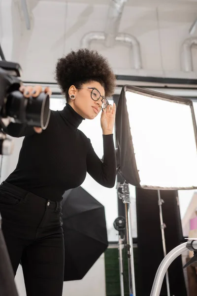 Elegante gestore di contenuti africani americani che regola gli occhiali vicino alla fotocamera digitale offuscata e al riflettore softbox in studio fotografico — Foto stock