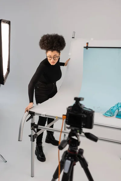 Afro-américain fabricant de contenu ajuster la table de tir avec des sandales à la mode près de l'appareil photo numérique flou dans le studio photo — Photo de stock