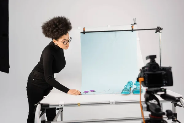 African american content producer looking at stylish sunglasses and sandals on shooting table near blurred digital camera in photo studio — Stock Photo