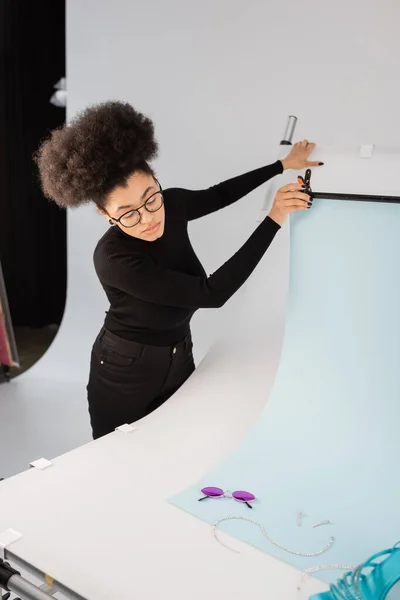 African american content maker adjusting background sheet near trendy sunglasses and jewelry on shooting table in photo studio — Stock Photo