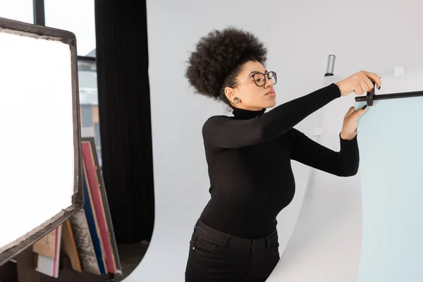 Produtor de conteúdo afro-americano em óculos instalando folha de fundo perto do refletor no estúdio de fotografia — Fotografia de Stock