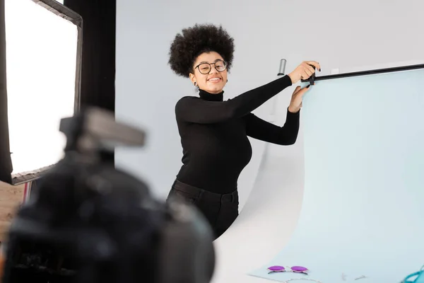 Happy african american content producer in eyeglasses installing background sheet in photo studio on blurred foreground — Stock Photo