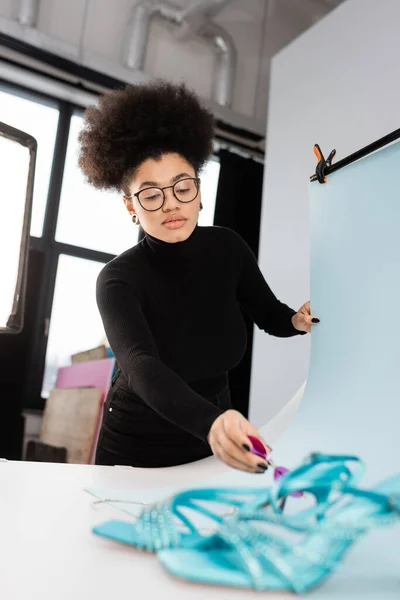 African american content producer placing sunglasses near blurred sandals on shooting table in photo studio — Stock Photo