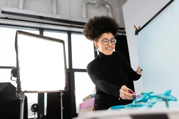 Cheerful african american content maker in eyeglasses looking at camera near reflector and shooting table in photo studio — Stock Photo