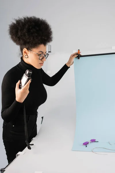 Produtor de conteúdo americano africano de pé com medidor de luz perto de folha de fundo branco e óculos de sol na moda na mesa de tiro — Fotografia de Stock