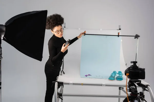 African american content producer looking at light meter near reflector and shooting table with trendy footwear and sunglasses in photo studio — Stock Photo