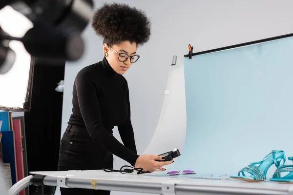 Fabricante de contenido afroamericano utilizando medidor de luz cerca de gafas de sol de moda y sandalias en la mesa de tiro en el estudio de fotos - foto de stock
