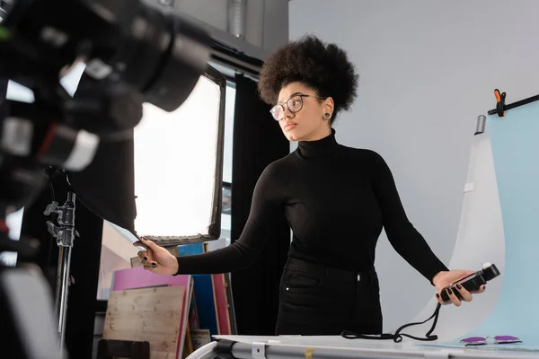 Produtor de conteúdo afro-americano em óculos segurando medidor de exposição perto da mesa de tiro no estúdio de fotos — Fotografia de Stock