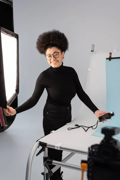 Produtor de conteúdo americano africano alegre com medidor de exposição olhando para a câmera perto de holofotes e mesa de tiro no estúdio de fotos — Fotografia de Stock