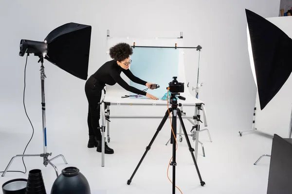 Produtor de conteúdo americano africano com medidor de exposição perto de mesa de tiro e equipamentos de iluminação no estúdio de fotografia moderna — Fotografia de Stock
