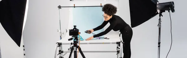 African american content producer with exposure meter near shooting table and reflectors with digital camera in photo studio, banner — Stock Photo