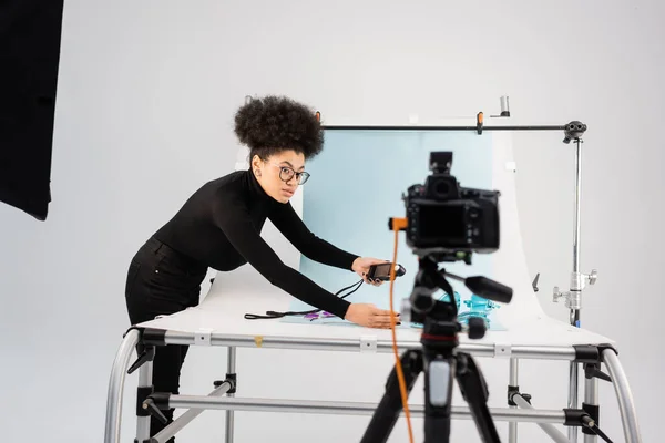 Créateur de contenu afro-américain regardant un appareil photo numérique flou tout en tenant un compteur d'exposition près de la table de tir dans un studio photo moderne — Photo de stock