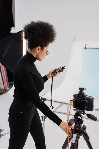 Gerente de contenido afroamericano mirando el medidor de exposición cerca de la cámara digital y la mesa de fotos en el estudio de fotografía - foto de stock