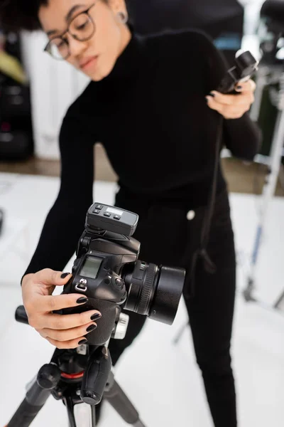 Productor de contenido afroamericano borroso sosteniendo el medidor de exposición y ajustando la cámara digital en el estudio de fotografía - foto de stock