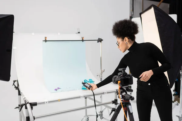 Gerente de conteúdo afro-americano segurando medidor de exposição perto de sandálias e óculos de sol na mesa de tiro no estúdio de fotos — Fotografia de Stock