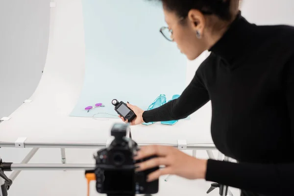 Vista lateral del productor de contenido afroamericano que sostiene el medidor de exposición entre la cámara digital borrosa y la mesa de fotos en el estudio de fotografía - foto de stock
