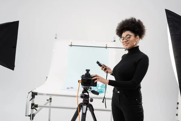 Fabricante de conteúdo americano africano feliz e elegante com medidor de exposição perto de câmera digital e mesa de tiro no estúdio moderno — Fotografia de Stock