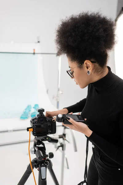 Side view of african american content producer with exposure meter adjusting modern digital camera in photo studio — Stock Photo