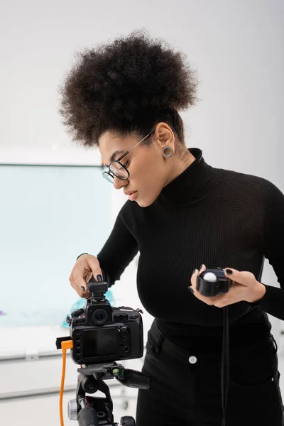 Elegante gerenciador de conteúdo afro-americano segurando medidor de exposição e ajustando câmera digital no estúdio de fotos — Fotografia de Stock