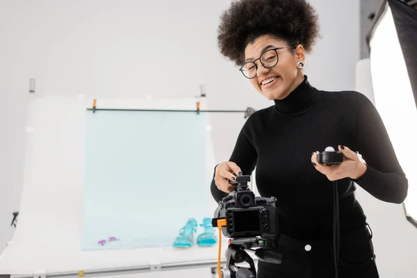 Fabricante de contenido afroamericano alegre con medidor de exposición sonriendo cerca de la cámara digital en el estudio moderno - foto de stock