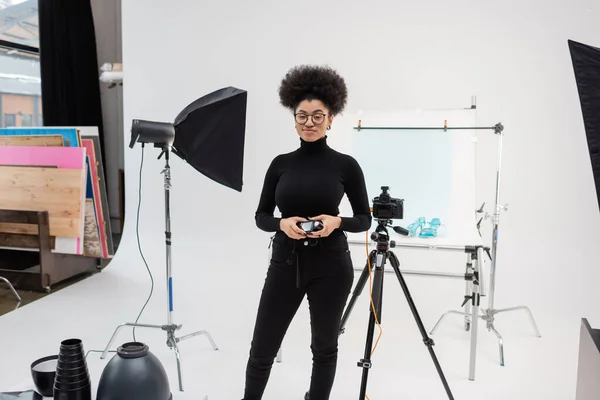 Усміхнений афроамериканський виробник контенту стоїть з вимірювачем експозиції біля цифрової камери та освітлювального обладнання в сучасній фотостудії — стокове фото