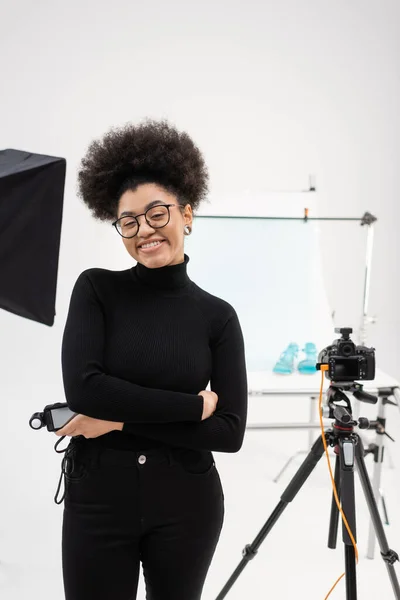 Productor de contenido afroamericano satisfecho con medidor de exposición sonriendo cerca de la cámara digital en el estudio de fotos - foto de stock