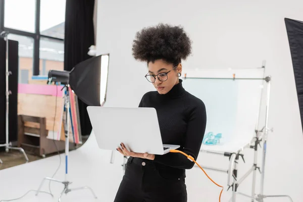 Productor de contenido afroamericano en anteojos mirando portátil cerca de reflector softbox y mesa de tiro en estudio de fotos — Stock Photo