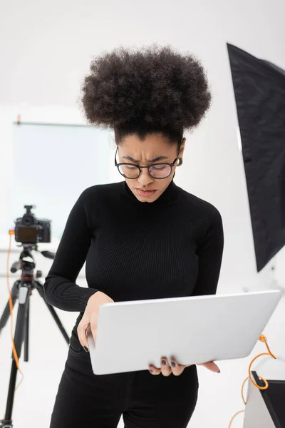 Disgustado y frunciendo el ceño productor de contenido afroamericano utilizando el ordenador portátil cerca de la cámara digital borrosa en el estudio de fotos - foto de stock