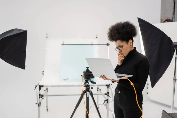 Thoughtful african american content manager looking at laptop near spotlights and digital camera in modern photo studio — Stock Photo