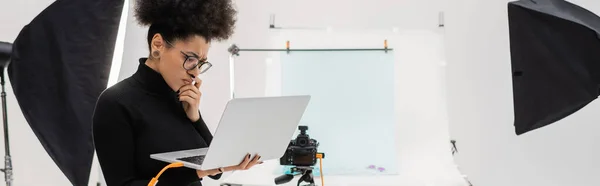 Serio produttore di contenuti afro-americani guardando il laptop mentre pensa vicino ai riflettori e alla fotocamera digitale in studio fotografico, banner — Foto stock