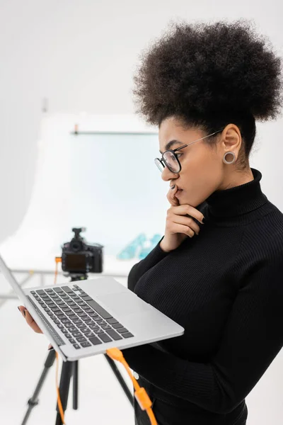 Vista lateral do pensativo fabricante de conteúdo afro-americano olhando para o laptop no estúdio de fotos borrado — Fotografia de Stock