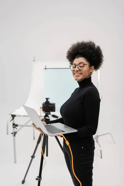 Produtor de conteúdo americano africano feliz em óculos segurando laptop e olhando para a câmera no estúdio de fotos — Fotografia de Stock