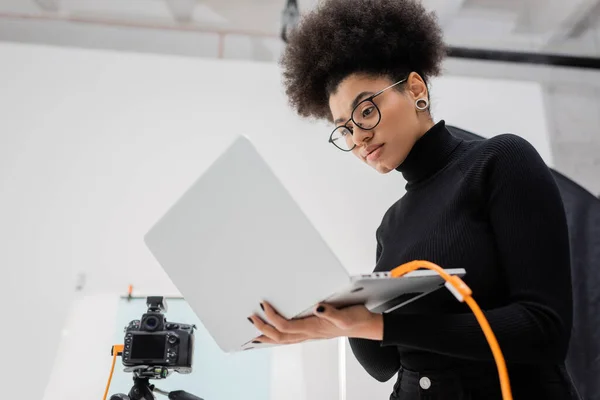 Visão de baixo ângulo de produtor de conteúdo americano africano elegante em óculos de pé com laptop perto de câmera digital no estúdio de fotos — Fotografia de Stock
