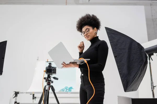 Fabricante de conteúdo afro-americano com laptop sorrindo perto de softbox e câmera digital no estúdio de fotos contemporâneo — Fotografia de Stock