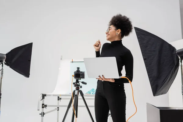 Fabricante de contenido afroamericano alegre con portátil mirando hacia otro lado cerca de softboxes y cámara digital en el estudio de fotos moderno - foto de stock