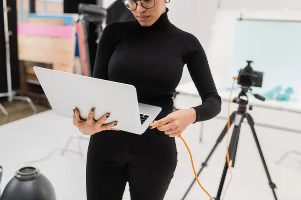 African american content producer connecting cable to laptop near blurred digital camera in photo studio — Stock Photo
