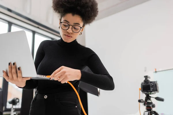 African american content manager in eyeglasses and black turtleneck connecting cable to laptop in photo studio — Stock Photo