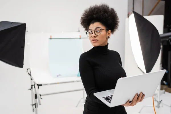 African american content manager with laptop looking away near spotlights and blurred shooting table in photo studio — Stock Photo