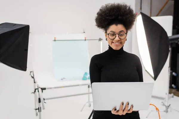 Produtor de conteúdo americano africano feliz com laptop sorrindo para a câmera perto de refletores e mesa de tiro no estúdio de fotos — Fotografia de Stock