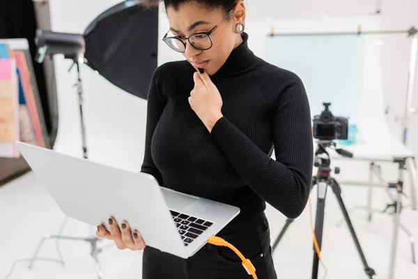 Pensativo Africano americano fabricante de conteúdo em óculos olhando para laptop perto de câmera digital borrada no estúdio de fotos — Fotografia de Stock