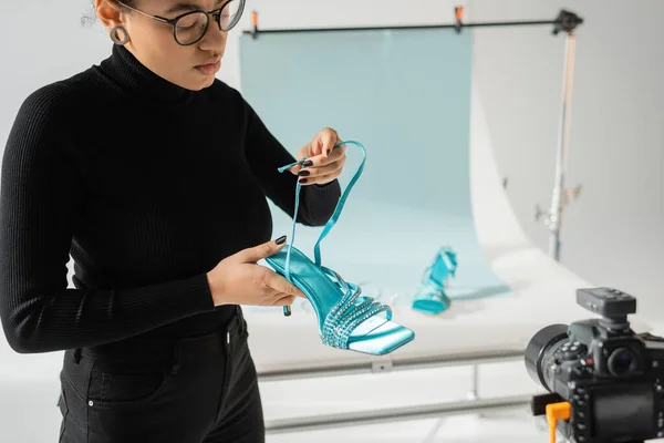 Afrikanisch-amerikanischer Content-Hersteller mit Brille hält trendige Sandale in der Nähe verschwommener Digitalkamera im Fotostudio — Stockfoto